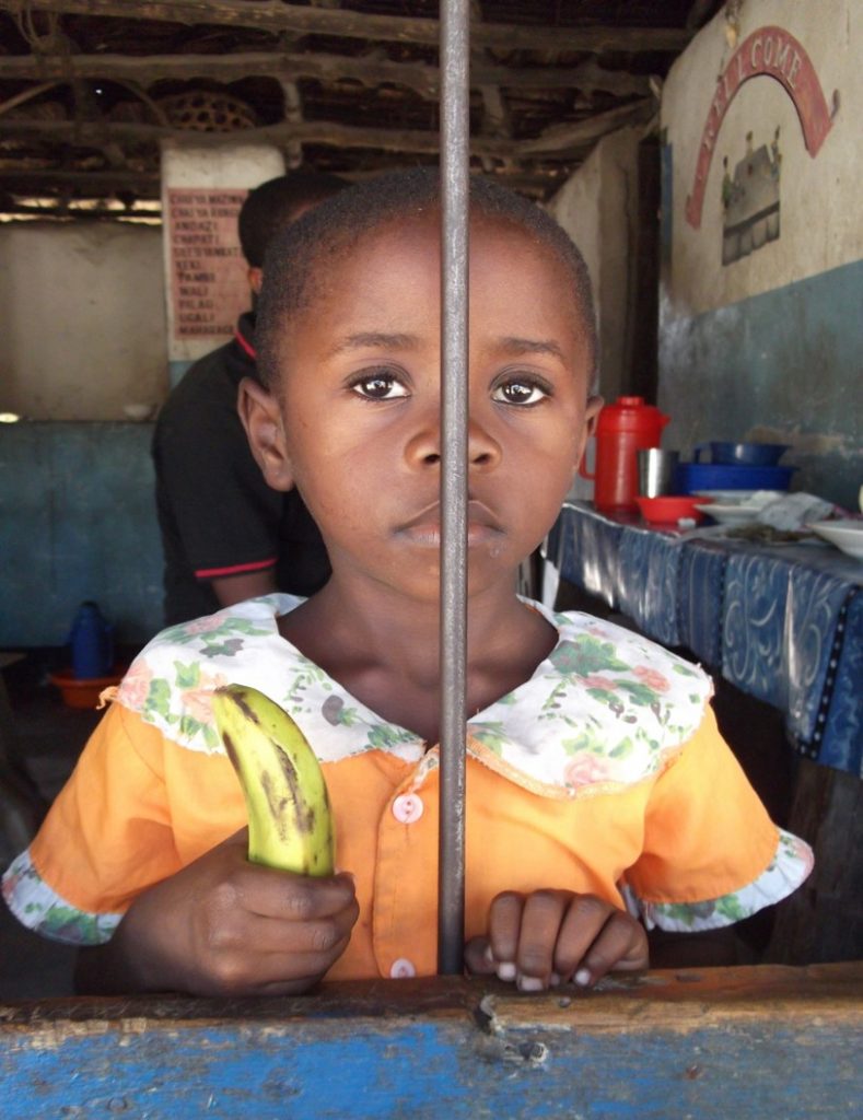 Small child holding a banana behind a bar